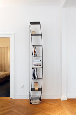 Gespanntes Regal von Moormann, gefüllt mit Büchern und Zeitungen, vor weißer Wand und Parkettboden.